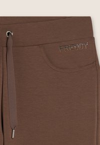 Pantaloni della tuta marroni con una texture morbida, dotati di tasca laterale e vita con cordoncino. Il marchio "FREDDY" è decorato con strass.