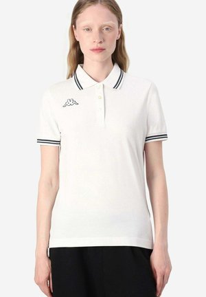 Giovane donna che indossa una polo bianca con colletto e maniche a righe nere, con un piccolo logo sul petto a sinistra.