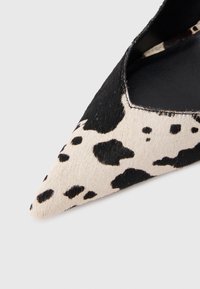 Zapato de mujer con punta puntiaguda, patrón de piel de vaca en blanco y negro, y costuras visibles a lo largo del borde sobre un fondo blanco.