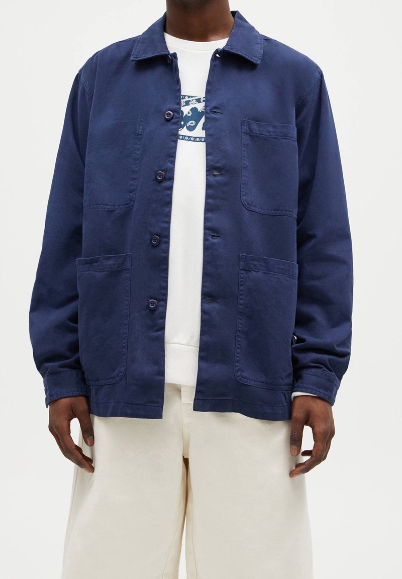 Mörkblå button-up jacka i bomull med tre framfickor, krage och en avslappnad passform, bärs över en vit sweatshirt och ljusa byxor.