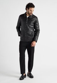 Zwarte leren jas met een kraag, voor-rits en een gladde textuur. Gdragen over zwarte pantalon en gecombineerd met zwarte instappers.