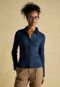 Navy blue, ribbed button-up shirt with a pointed collar and long sleeves, paired with tan plaid trousers. Plain buttons, fitted design.