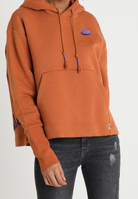 Person som bär en bränd orange huvtröja med blå broderade detaljer och svarta slitna jeans mot en enfärgad bakgrund.