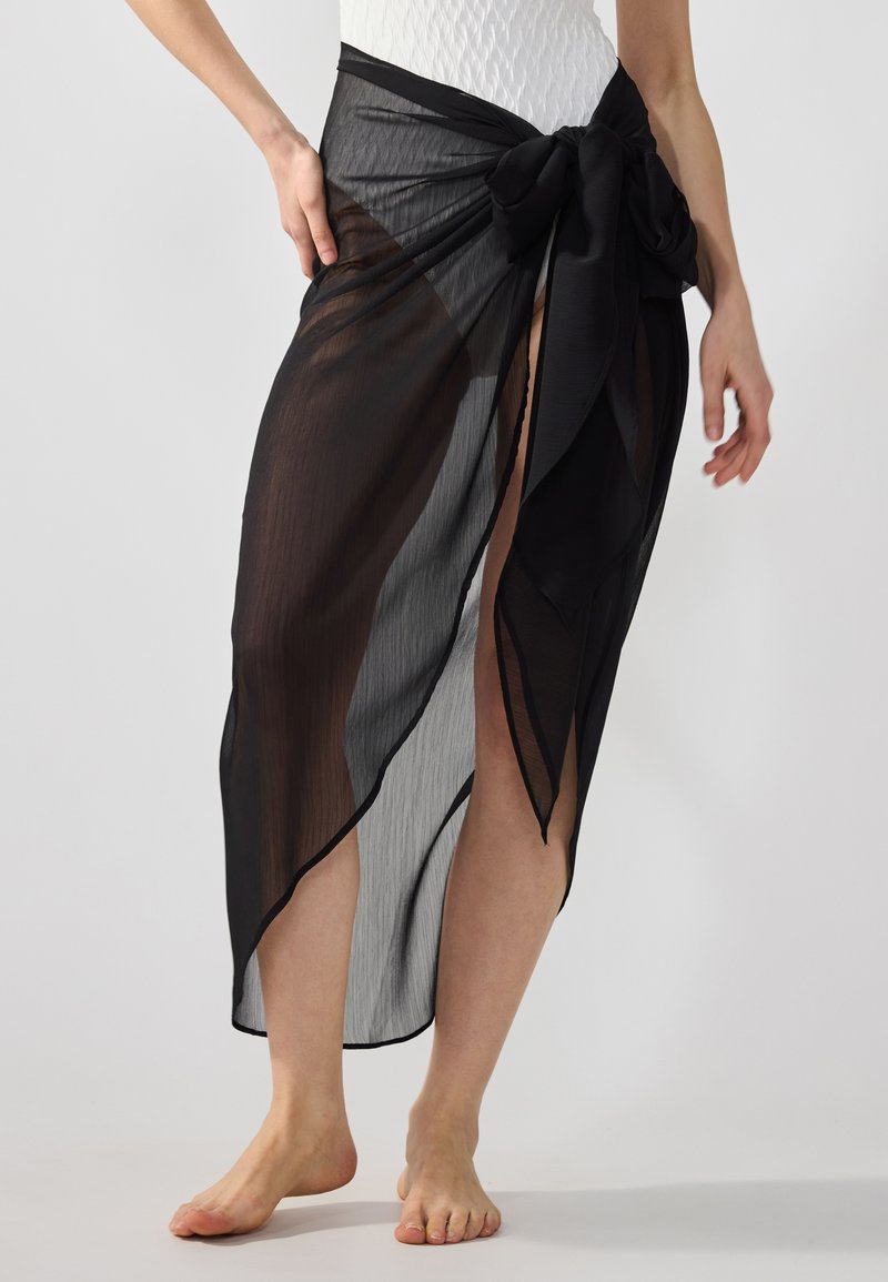 Jupe noire transparente à nouer à la taille, avec une texture légèrement translucide et une fente haute sur un côté.