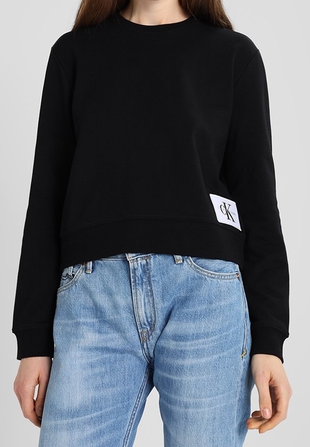 Osoba ubrana w czarną, długą bluzę crop top z naszywką z logo Calvin Klein oraz jasnoniebieskie, wysokogurtowe dżinsy.