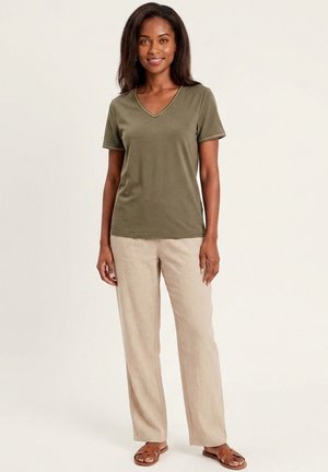Femme debout, portant un t-shirt vert olive à col en V et manches courtes, un pantalon large beige et des sandales marron, souriante devant un fond clair uni.