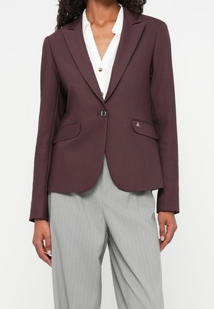 Kvinde iført en figursyet mørkelilla blazer over en hvid skjorte med knapper og lysegrå bukser med nålestriber mod en ensfarvet baggrund.