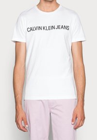 Bílý bavlněný tričko s nápisem "CALVIN KLEIN JEANS" v tučném černém písmu na prsou, krátké rukávy a kulatý výstřih.