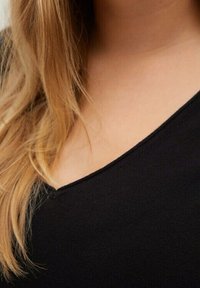 Czarna bluzka z dekoltem w serek o gładkiej strukturze, dopasowanym kroju i minimalnym przeszyciu. Długie, falujące blond włosy częściowo widoczne.