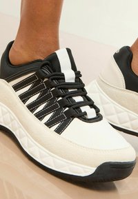 Sneaker cu un design alb și negru, având un superior texturat, șireturi elastice și o talpă cu model într-o formă joasă.