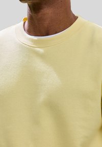 Sweat-shirt jaune clair en tissu doux, avec un col rond et une texture lisse. Des détails de coutures sont visibles près du col.