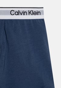Tamno plave bokserice s glatkom teksturom, s bijelim pojasem i crnim logotipom "Calvin Klein" te sivim naglascima.