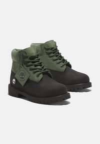 Stivali impermeabili neri e verde scuro con textures in suede, occhielli in metallo, lacci verdi e un’etichetta con un logo sul lato.