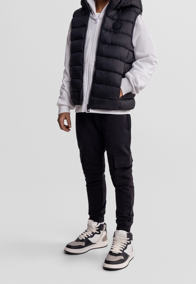Enfant portant une doudoune noire sur un sweat à capuche blanc, un jogger cargo noir et des baskets montantes noir et blanc, debout avec une main dans la poche.