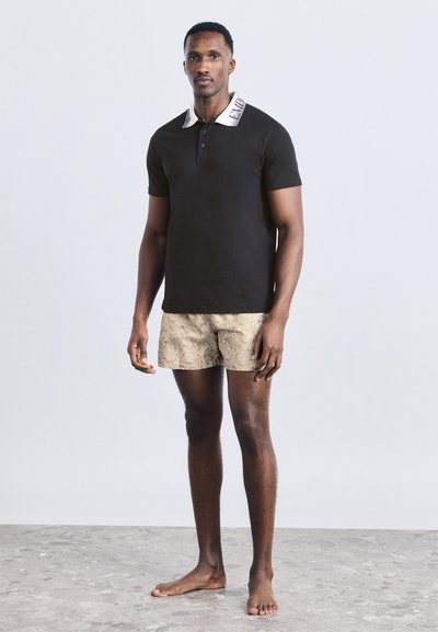 Άνδρας στέκεται ξυπόλητος, φορώντας κοντομάνικο μαύρο πόλο με λευκό γιακά και μπεζ σορτς με σχέδια, μπροστά σε απλό φόντο.