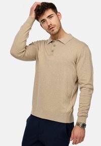 Polo beige à manches longues avec patte de boutonnage à trois boutons, poignets et ourlet côtelés. Porté avec un pantalon sombre et une montre au poignet.