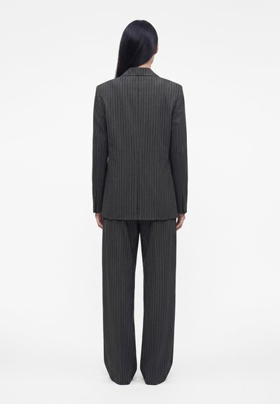 Personne aux longs cheveux lisses et foncés vue de dos, portant un costume à fines rayures gris foncé avec un pantalon ample et un blazer, sur un fond uni.