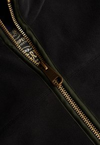 Zwarte fleece hoodie met een gouden ritssluiting, voorzien van een groene ritsafwerking. Binnenlabel toont "Superdry." Zachte textuur, zichtbare naden.