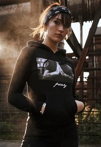 Sudadera negra con un gráfico de manos en oración y la palabra "reza". Presenta un bolsillo frontal y un diseño ajustado con capucha.