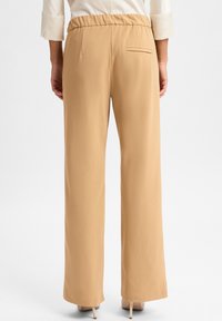 Beige weit geschnittene Hose mit elastischem Bund und einer einzigen Gesäßtasche, aus glattem Stoff, von hinten mit Absätzen präsentiert.