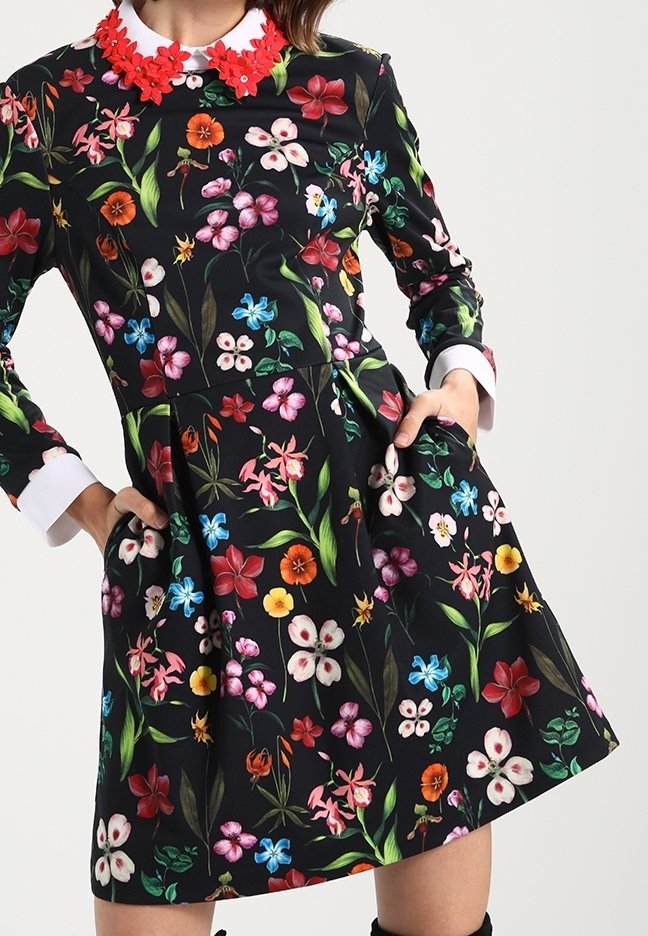 Femme portant une robe noire à manches longues avec des motifs floraux colorés, des poignets blancs et un col orné d'une fleur rouge, les mains dans les poches.