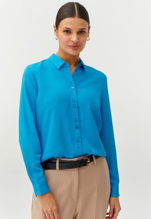 Camicia azzurra a maniche lunghe con colletto, realizzata in tessuto liscio, abbinata a pantaloni beige e una cintura nera.