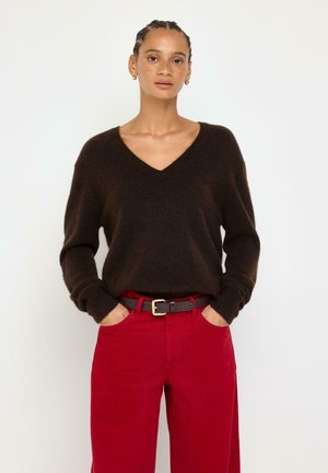 Donna con trecce che indossa un maglione nero a V e pantaloni rossi con cintura nera, in piedi con le mani in tasca davanti a uno sfondo bianco.