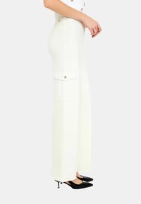 Pantaloni bianchi a gamba larga in tessuto texturizzato, dotati di una tasca laterale con chiusura a button e indossati con tacchi neri a punta.