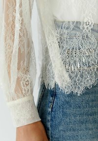 Kanten blouse met doorschijnende mouwen, complexe bloemenpatronen, een gegolfde zoom en een crèmekleurige tint, gecombineerd met blauwe denim jeans.
