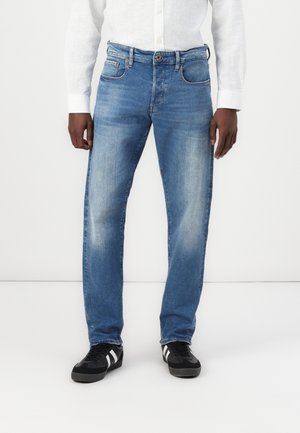 Homme portant un jean bleu délavé, une chemise blanche boutonnée et des baskets noires avec des rayures blanches, debout devant un fond blanc uni.