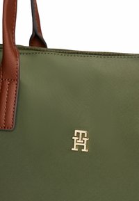 Groene stoffen tas met bruine leren handvatten en een gouden "TH" logo-embleem nabij de bovenste rits.