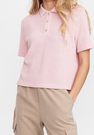 Femme portant un polo rose clair texturé à manches courtes et un pantalon beige taille haute, main dans la poche, visible des épaules aux hanches.