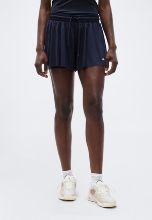 Frau trägt marineblaue Sportshorts, weiße Knöchelsocken und weiße Turnschuhe, steht vor einem schlichten hellen Hintergrund.