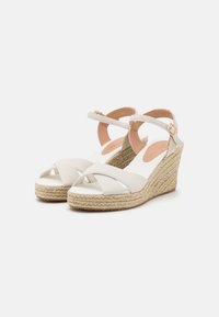 Wedge sandals featuring a beige canvas upper with crisscross straps, espadrille-style jute sole, and adjustable ankle strap with gold hardware.
