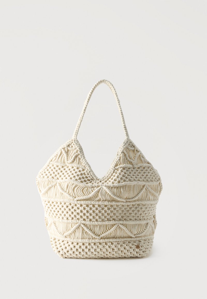 Τσάντα tote μακραμέ σε χρώμα κρέμα με περίτεχνα υφαντά σχέδια και μία χειρολαβή σε σχήμα θηλιάς σε απλό φόντο.