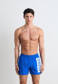 Shorts de bain bleus en tissu léger, dotés d'une taille élastique et d'un grand logo blanc "HUGO" sur le côté.