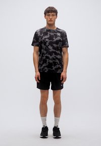 Μαύρο κοντομάνικο πουκάμισο με καμουφλάζ σχέδιο και στρογγυλή λαιμόκοψη, σε συνδυασμό με μαύρο σορτς. Φορεμένο με λευκές κάλτσες και μαύρα αθλητικά παπούτσια.