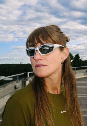Mujer con cabello largo y castaño que lleva gafas de sol deportivas blancas y una blusa verde oliva, de pie al aire libre con un cielo nublado de fondo.