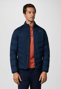 Námořnická modrá prošívaná bunda s límcem, zapínáním na zip a detailem loga, nošená přes červenou košili. Hladká textura, lehký design.