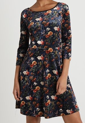 Femme portant une robe en velours bleu marine avec un motif floral multicolore et des manches trois-quarts, debout devant un fond uni.