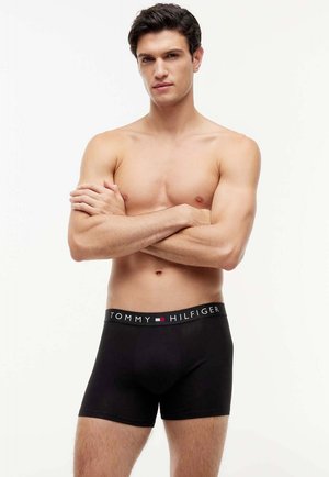 Uomo senza maglietta con le braccia incrociate che indossa boxer neri Tommy Hilfiger su uno sfondo bianco uniforme.