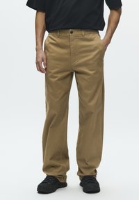 Pantalon en coton kaki avec une coupe droite, poches avant et une fermeture à un bouton. Porté avec des baskets noires, affichant un style décontracté.