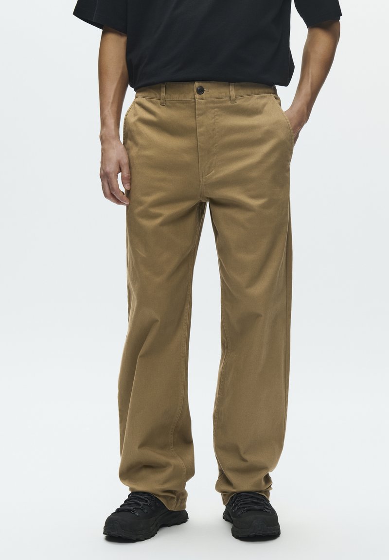 Pantalon en coton kaki avec une coupe droite, poches avant et une fermeture à un bouton. Porté avec des baskets noires, affichant un style décontracté.