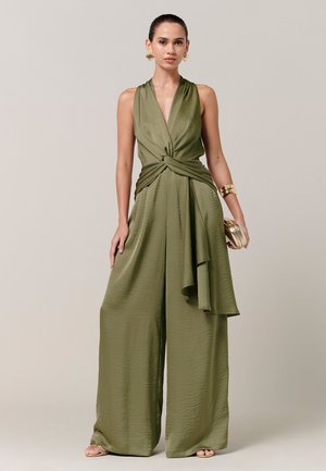 Kvinde iført olivengrøn ærmeløs buksedragt med vide ben og knudedetalje, guldbøjlearmbånd, øreringe og holder en lille gylden clutch.