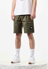 Shorts cargo vert olive avec plusieurs poches, une étiquette noire, et une coupe décontractée. Portés avec des chaussures de sport blanches et des chaussettes blanches.