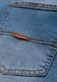 Tissu en denim bleu clair avec une texture visible, présentant une étiquette en cuir marron marquée "ombre dept.", cousue avec un fil contrastant.