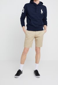 Muž oblečený v tmavě modré mikině s bílým logem polokošile, béžových šortkách, bílých ponožkách a černých teniskách, stojící s rukama v kapsách.