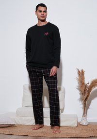 Cămașă neagră cu mânecă lungă, având o mică grafică cu un cerb roșu, purtată cu pantaloni de pijama în carouri negre și roșii. Este îmbrăcată cu picioarele goale, stând pe un covor.