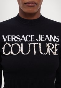 Pull en tricot noir avec un col montant, arborant "VERSACE JEANS" en blanc et "COUTURE" orné de perles et de strass.