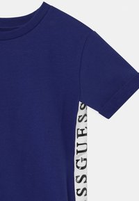 Himmelblaues Kurzarmhemd mit umgeschlagenen Manschetten und weißen Seitestreifen, auf denen vertikal schwarzer "GUESS"-Text steht.